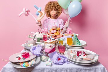 Unhappy curly haired woman celebrates birthday alone stands near festive table with desserts holds inflated balloons isolated over pink background. People celebration and special occasion concept