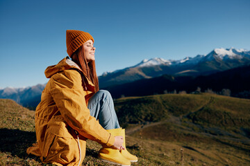 Woman sitting on a hill on the grass rest smile with teeth looking at the mountains in the snow in the autumn in a yellow raincoat and jeans happy sunset hiking trip, freedom lifestyle 