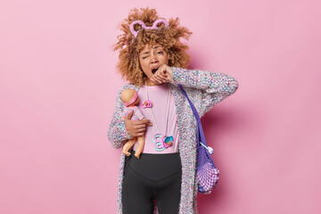 Tired pregnant young woman yawns and covers mouth has leaked makeup wears warm jumper and leggings holds doll going to become mother of daughter carries net bag isolated over pink background