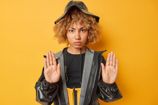 Serious woman with curly hair shows stop denial gesture disapproval sign rejects and prohibits something wears black leather hat and raincoat isolated over yellow background. Body language concept