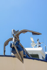 A heavy sturdy steal anchor of a large docked fishing boat hangs over the bow of the vessel with the bridge of the trawler in view- Ready for work. Vertically set against a cloudless bright blue sky.