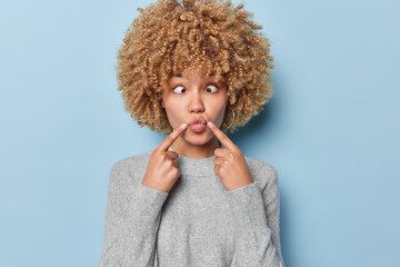 Horizontal shot of funny woman goes crazy makes grimace pours lips and crosses eyes wears earrings and grey casual jumper isolated over blue background tries to make friends laugh. Human expressions