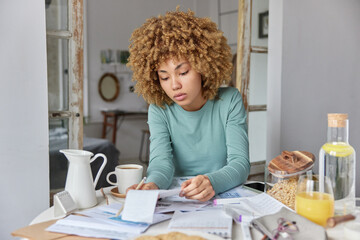Beautiful woman studies financial bills dressed in casual jumper sits at table poses against cozy interior plans money savings checks reports does paperwork. Planning budget and managing expenses