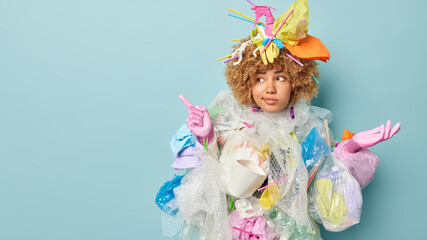 Photo of serious woman wears gloves surrounded by plastic wastes cleans Earth from rubbish points index finger left on blank space demonstrates place for your promotion isolated over blue background