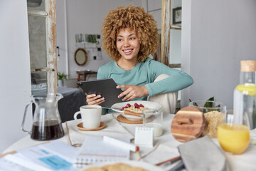 Photo of beautiful curly woman uses tablet for scrolling newsfeed eats cereals for breakfast poses...