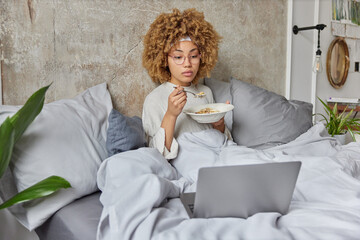 Serious woman eats oatmeal for breakfast relaxes in bed watches intersting film via laptop computer...