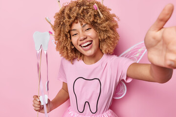 Horizontal shot of positive woman pretends being tooth fairy smiles broadly holds wand tells how to...