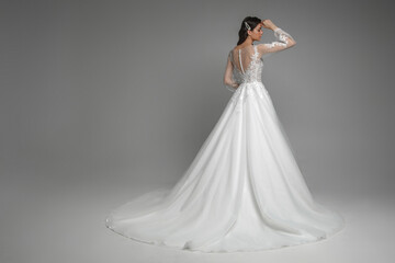 Back view of the beautiful brunette bride wearing modern white wedding dress with pattern and pearls on the back. Gorgeous female posing with hands on a grey isolated background.