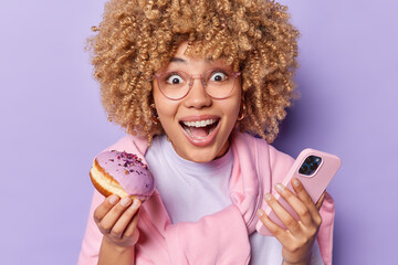 Happy surprised curly haired woman looks gladfully at camera holds delicious doughnut and mobile...