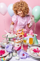 Photo of cheerful curly haired woman wears checkered dress silk gloves tries new dessert stands near table with various sweet food on birthday surrounded by inflated balloons. Festive event concept