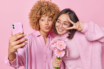 Horizontal shot of two mixed race women pose for making selfie make peace gesture hold gerbera flowers dressed in pink clothes stand against rosy wall. Sisters make photo for social networks