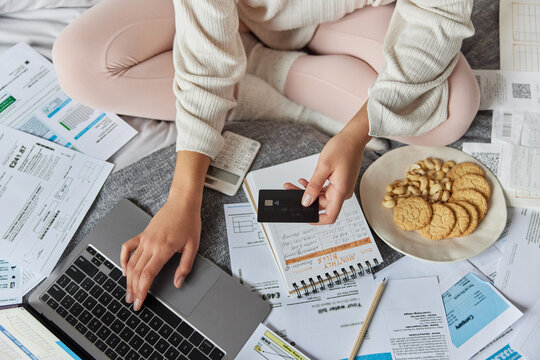 Unknown woman enters data in computer application holds credit banking card reviews paperwork documents manages financial papers sits crossed legs on bed eats snack dressed in domestic clothes