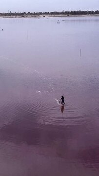 Vertical video of Man stiring water in middle of pink lake with boat full of salt