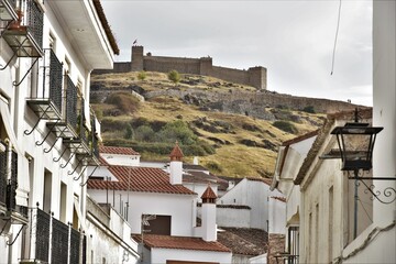 Rampart of Castillo de Aracena fortress atop a barren hill covered with sparse yellow brownish grasses and boulders seen thru a white lane of Aracena country township, Aracena, Spain