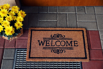Door mat with word Welcome and beautiful flowers near entrance