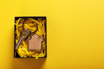 Key with trinket in shape of house and gift box on yellow background, top view with space for text. Housewarming party