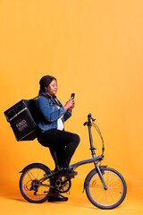 Restaurant employee standing on bike in studio with yellow background while checking customer adreess on phone before start delivery takeaway meal. Pizzeria courier carrying take out backpack