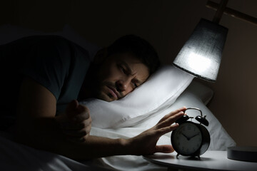 Sleepy man turning off alarm clock on nightstand in morning