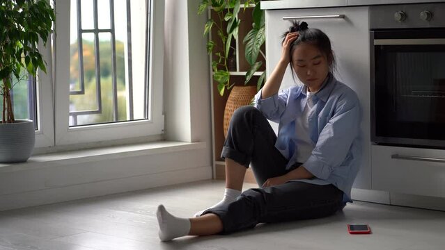 Upset depressed Asian girl suffering from breakup, heartbroken frustrated woman sits on floor in kitchen looking at smartphone waiting for call or message from ex boyfriend. Codependent relationships
