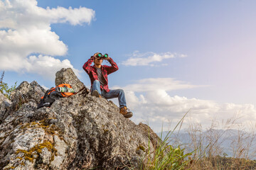 Male hiker with backpack holding binoculars sitting on top of the rock mountain