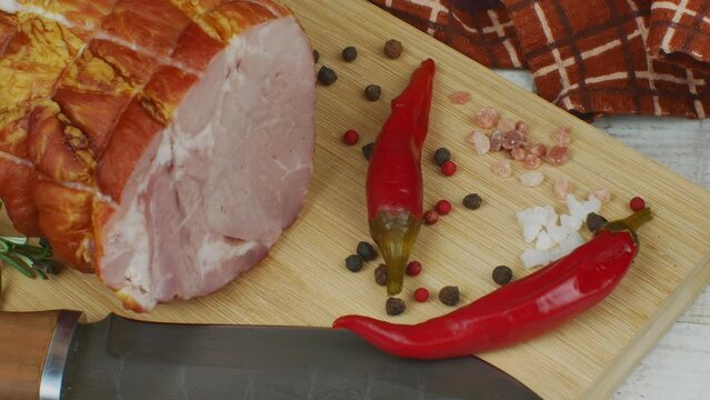 A juicy piece of pork ham on a wooden cutting board, next to it are a carving knife, multi-colored allspice peas, red hot peppers, parsley, dill and basil. The concept of delicious meat products