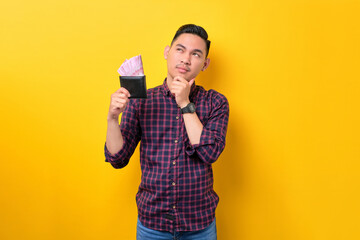 Pensive young Asian man holding wallet full of money banknotes and looking at copy space isolated...