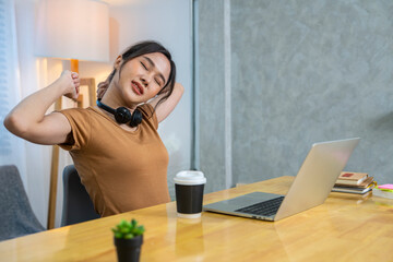 Asian woman working at home wearing headphones typing in homework reports until sleepy and feeling shoulder pain, neck pain, having to stretch arms like office syndrome