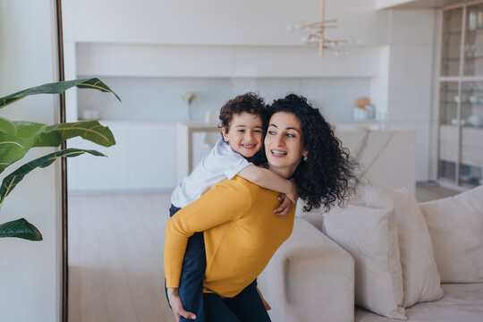 Young Spanish curly woman having fun with son, rolls him on back looking aside. Babysitter in yellow sweater and green pants entertains little cheerful child at home. Elder sister plays with brother.