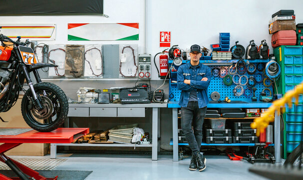 Portrait of mechanic man wearing jeans and cap standing in front of workbench in motorbike workshop