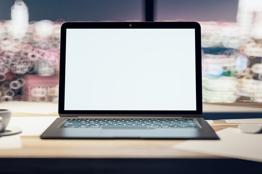 Front view on blank white modern laptop monitor with space for your logo or text on light wooden surface on blurred night city view from window background, close up. 3D rendering, mock up