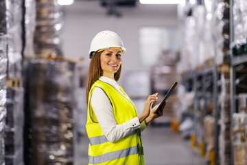 Portrait of a smiling female controller standing in factory warehouse and using tablet.