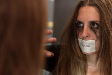 a crying woman looking at herself in the mirror with her mouth covered with a tape with the text...