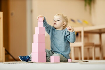 A focused child is putting blocks and making tower at montessori kindergarten. Learning through the...