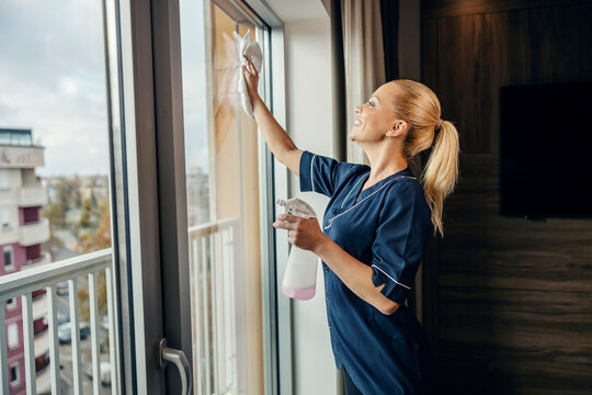 A Happy Maid Is Cleaning Windows In Hotel Room With Detergent And Cloth.