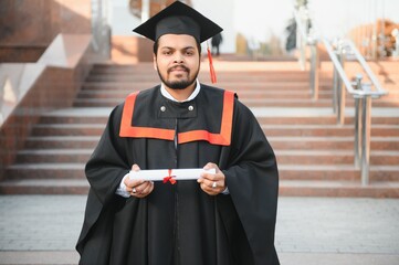 Portrait of handsome indian graduate student in graduation glow with diploma.