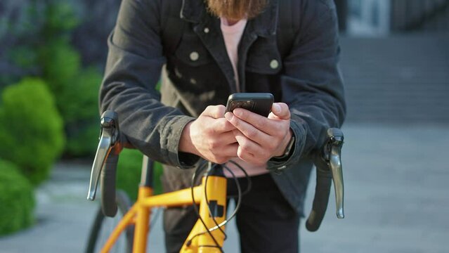Close up of man with beard wearing casual clothes, standing with bicycle, leaning. Maale holding smartphone, using, scrolling, texting, messaging. Concept of modern urban living.