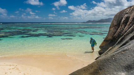 A man walks on a sandy beach, on the water. Backpacks on the shoulders. The turquoise ocean is calm. Clouds in the blue sky. In the foreground are the slopes of coastal granite rocks. Seychelles.