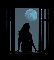 full moon in the night dark blue sky. Black silhouette of  magical witch woman looking out of an...