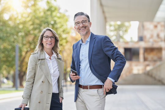 Cheerful businesswoman and businessman with smart phone standing on footpath - Powered by Adobe