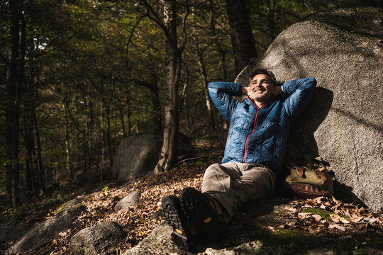 Happy Mature Man With Hands Behind Head Relaxing By Rock In Forest