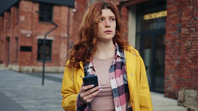 Good-looking young woman lost in city. Redhead girl holding smartphone, looking at address on buildings, using navigator. Digital technologies. Internet connection. Unknown city. Tourism concept