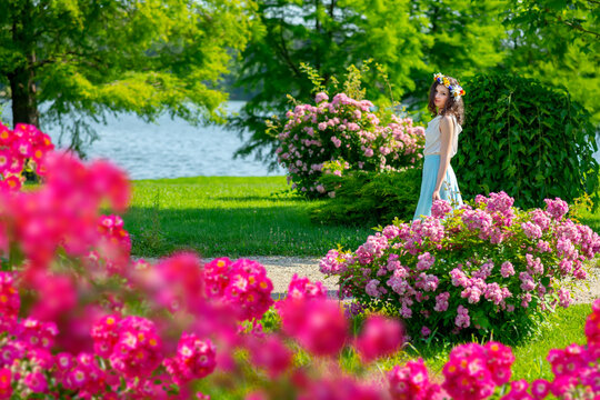 Outdoor Portrait Of Young Happy Smiling Fantasy Woman Walking Among Flowers. Young Beautiful Goddess Enjoys Spring Nature In Bright Sunlight. Fairy Tale Image Art Photo With Pink Roses Frame