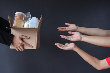 Hand giving box of various foods to many hands on dark gray background. Donation and charity concept