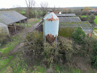 Large, metal grain feed silo showing a  poor state of repair. Surrounding farm buildings are seen belonging to a British dairy farm.
