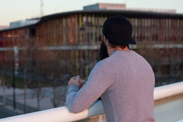 hombre caucásico con barba y gorra, apoyado en un puente mirando al horizonte en una ciudad urbana
