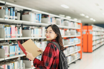 Female student in casual style is opening books and smiling to the camera against blurred library background.