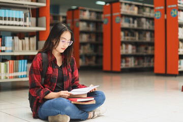 College female student wearing casual style and backpack is sitting and reading a book against blurred library background.