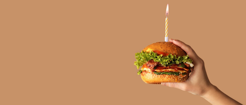 Female hand holding tasty burger with burning Birthday candle on brown background with space for text