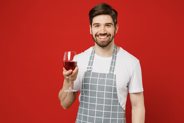 Young smiling sommelier fun happy male housewife housekeeper chef cook baker man wear grey apron hold in hand glass of wine look camera isolated on plain red background studio. Cooking food concept.