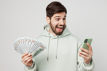 Young excited caucasian man wear mint hoody use mobile cell phone hold in hand fan of cash money in...